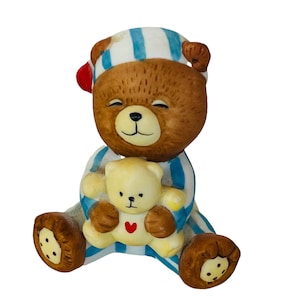 Puede incluir: Un oso de peluche marrón que lleva un pijama a rayas azules y blancas está sosteniendo un oso de peluche amarillo más pequeño. El oso más grande tiene un gorro rojo y una expresión feliz.