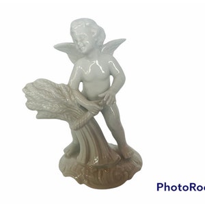 Puede incluir: Una figurita de querubín de cerámica blanca que sostiene una gavilla de trigo. El querubín tiene alas y está de pie sobre una base con un diseño de espiral marrón y blanco.