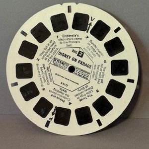 Könnte beinhalten: Eine cremefarbene Vintage-View-Master-Rolle mit Szenen aus Disneys "Disney on Parade". Die kreisförmige Rolle hat mehrere kleine rechteckige Fenster mit Bildern und Text, die Szenen aus dem Film beschreiben, wie Cinderella und Dumbo.