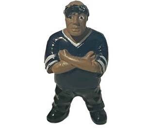 May include: A small plastic figurine of a man with brown skin, black hair, and a black and white striped shirt. He is standing with his arms crossed and has a serious expression on his face.