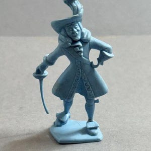 May include: A light blue figurine of a man in historical attire, holding a sword. The figure wears a hat with a feather plume and a long coat. The figurine stands on a square base.
