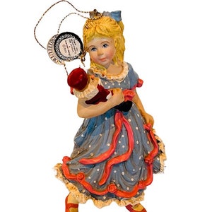 May include: A porcelain figurine ornament of a girl in a blue and white polka dot dress holding a red and black toy soldier. The girl has blonde hair and is wearing red shoes.