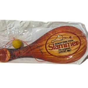 Puede incluir: Un Tootsietoy Slammer vintage, el "Wacky Bat" original, con una paleta de madera y una pelota amarilla unida por una cuerda. La paleta es marrón con una etiqueta roja y blanca que dice "Tootsietoy Slammer the original "Wacky Bat""