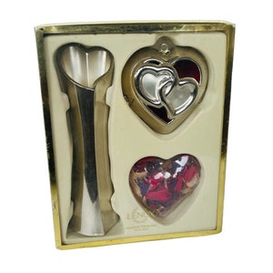 Pode incluir: Um conjunto de presente da Lenox com um vaso prateado em forma de coração, um ornamento prateado em forma de coração com dois corações entrelaçados e um recipiente em forma de coração cheio de potpourri vermelho e branco.