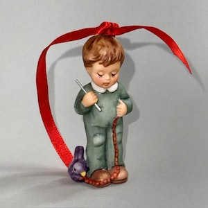May include: A ceramic figurine of a young child in a green outfit, holding a thin silver rod and a string of beads. A purple bird sits at the child's feet. The figurine is suspended by a red ribbon, suitable for hanging as an ornament.