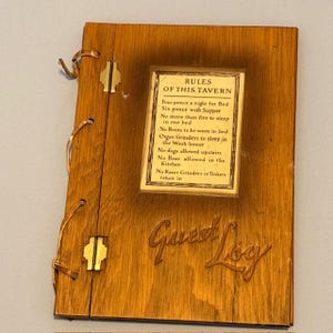 May include: Wooden guest log with the words "Guest Log" carved into the wood. A smaller rectangular sign is affixed to the log, displaying rules of the tavern. The log is bound with a leather cord and has metal hinges.