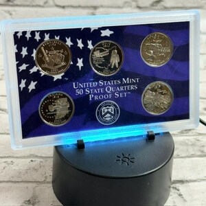Pode incluir: Um conjunto de moedas United States Mint 50 State Quarters Proof Set numa caixa retangular de plástico transparente. O conjunto apresenta cinco moedas de 2002 do Tennessee, Ohio, Louisiana, Indiana e Mississippi sobre um fundo de bandeira.