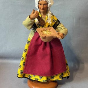 Puede incluir: Figura artesanal de una mujer con vestimenta tradicional. Lleva un gorro blanco, un vestido amarillo con estampado floral, un delantal rojo y una camisa verde de manga larga. La figura sostiene una herramienta de madera y una bolsa, posiblemente representando una escena de cocina o preparación de alimentos.