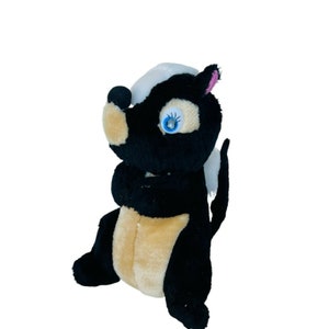 Può includere: Un puzzolo di peluche nero e bianco con il naso rosa e gli occhi blu. Il puzzolo è seduto con le braccia incrociate e ha una striscia bianca sulla schiena.