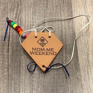 May include: A brown leather patch with the text "MOM-ME WEEKEND" and a Girl Scout logo. The patch is attached to a white cord with a rainbow of colorful beads and blue strings.