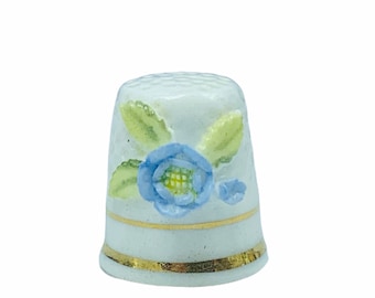 May include: White porcelain thimble with a blue and green floral design. The thimble has a gold trim around the top and bottom.
