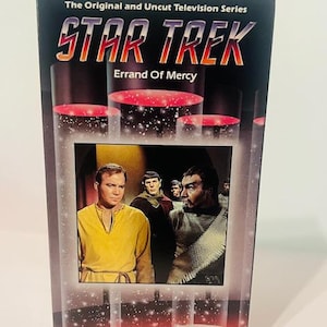 Puede incluir: Una cubierta de cinta VHS para el episodio de la serie de televisión Star Trek "Errand of Mercy". La cubierta presenta un fondo negro con estrellas blancas y un borde rojo. El título "Star Trek" está en letras blancas grandes con un contorno rojo. El título del episodio "Errand of Mercy" está en letras blancas más pequeñas debajo del título principal. La cubierta también presenta una escena del episodio con el Capitán Kirk y otros personajes.