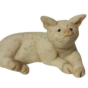 Peut inclure: Une petite figurine de cochon en céramique blanche couchée sur le côté, la tête tournée vers le côté. Le cochon a une texture mouchetée et une expression légèrement souriante.