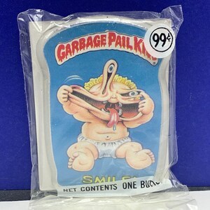 May include: A Garbage Pail Kids trading card featuring a cartoon baby with three eyes, a large mouth, and a long tongue. The card is blue and white with the text "Garbage Pail Kids" and "99¢" printed on it.
