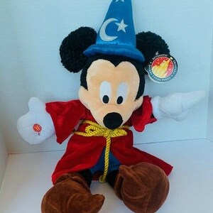 May include: A plush Mickey Mouse wearing a red robe and a blue wizard hat with a crescent moon and star. The plush toy has brown shoes and is holding a white glove in one hand. The tag reads "Authentic Disney Theme Parks Original".