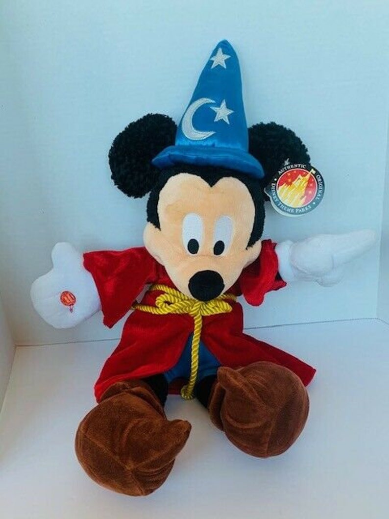 May include: A plush Mickey Mouse wearing a red robe and a blue wizard hat with a crescent moon and star. The plush toy has brown shoes and is holding a white glove in one hand. The tag reads "Authentic Disney Theme Parks Original".