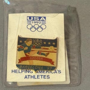 May include: A USA 1992 Olympic pin featuring a soccer player kicking a ball. The pin has a gold background with blue and red accents. Text includes "USA 1992", "Soccer", and "Barcelona '92". The pin is on a white card.