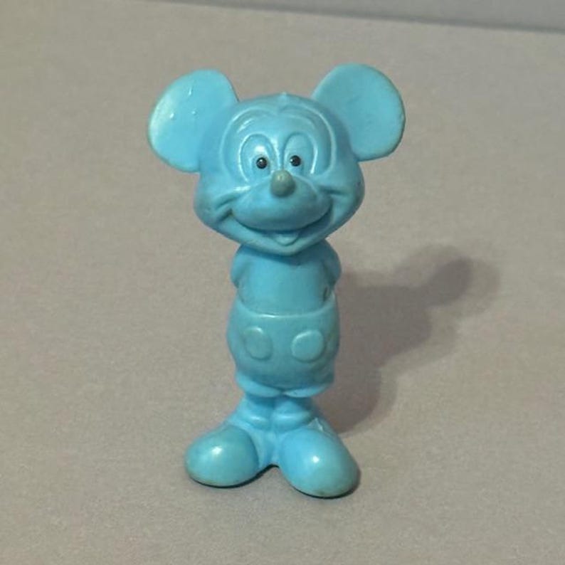 Pu&ograve; includere: Una statuetta di plastica blu di Topolino. La statuetta &egrave; in piedi con le braccia incrociate e ha un grande sorriso sul viso.