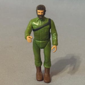 Op de afbeelding: Vintage actiefiguur van een man in een groen militair uniform met een zwarte band over de borst. De figuur heeft bruine laarzen en een donkere baard. De figuur staat op een grijs oppervlak.