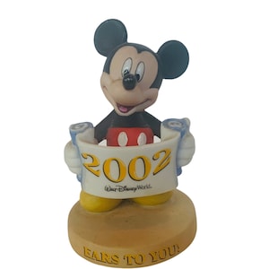 May include: A ceramic figurine of Mickey Mouse holding a scroll with the year 2002 and the text "Walt Disney World." The base of the figurine is brown and has the text "Ears to You!"