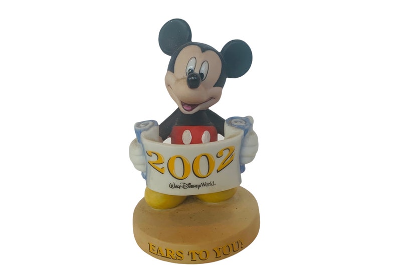 May include: A ceramic figurine of Mickey Mouse holding a scroll with the year 2002 and the text "Walt Disney World." The base of the figurine is brown and has the text "Ears to You!"