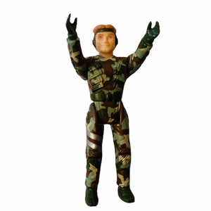 May include: A green and brown camouflage action figure with a light brown face and blonde hair. The figure is wearing a green and brown camouflage uniform with a black belt and a silver and black knee pad. The figure is standing with its arms raised above its head.