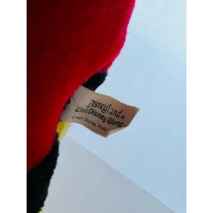 Puede incluir: Una etiqueta marr&oacute;n con el texto "Disneyland Walt Disney World" y un s&iacute;mbolo de copyright. La etiqueta est&aacute; unida a un juguete de peluche rojo, negro y amarillo.
