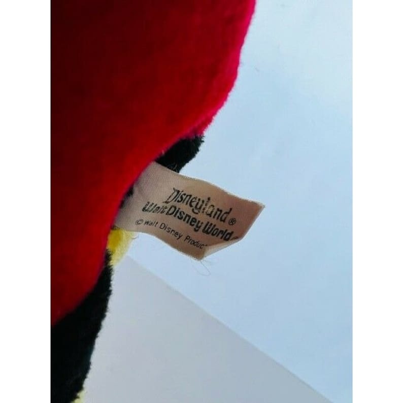 Puede incluir: Una etiqueta marr&oacute;n con el texto "Disneyland Walt Disney World" y un s&iacute;mbolo de copyright. La etiqueta est&aacute; unida a un juguete de peluche rojo, negro y amarillo.