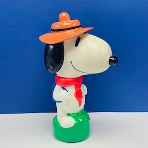 Puede incluir: Una figurita de plástico blanco de Snoopy con un sombrero marrón, una bufanda roja y una mochila azul. La figurita está de pie sobre una base verde.