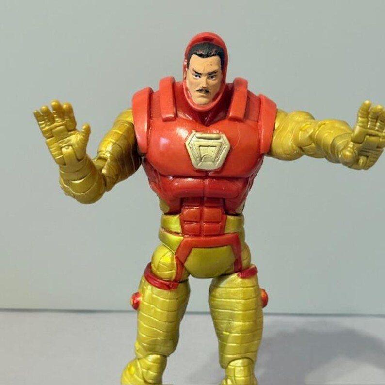 Op de afbeelding: Een rood en gouden Iron Man-actiefiguur met een snor. De figuur staat met zijn armen uitgestrekt, alsof hij in een verdedigende houding staat.