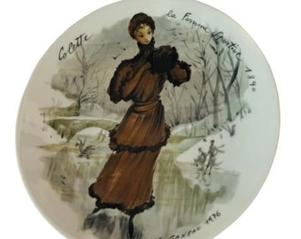 May include: A white ceramic plate with a painted image of a woman in a brown dress and hat walking in a snowy landscape. The plate is decorated with the text "La Femme Frontiere 1890" and "Colette FR Ganeau 1976".