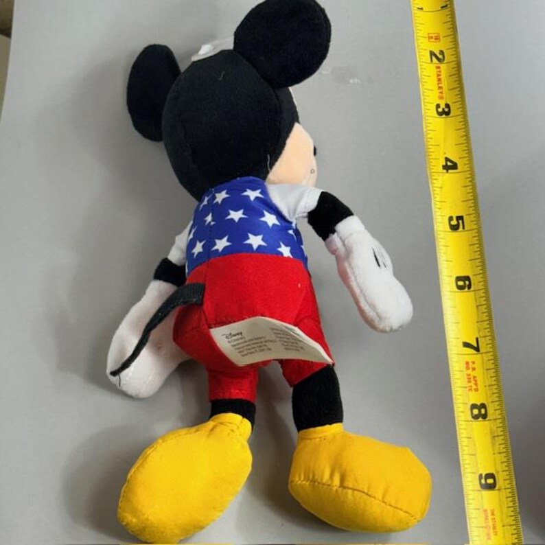 Op de afbeelding: Een Mickey Mouse pluche speelgoed, met een zwarte kop en oren, een wit gezicht en een patriottische outfit. De outfit bevat een blauwe top met witte sterren, een rode broek en gele schoenen. Het speelgoed is ongeveer 20 cm hoog.