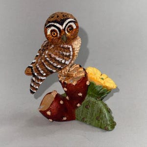 May include: A detailed figurine of an owl perched on a cactus. The owl has brown and white striped feathers, large orange eyes, and a detailed face. The cactus features a red fruit, a yellow flower, and green leaves.