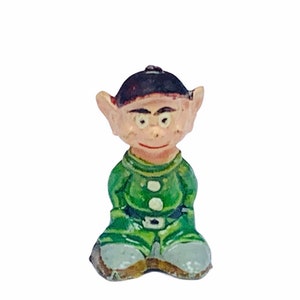May include: A small, green-clad figurine of a gnome with a black hat and a friendly expression. The gnome is sitting with its legs crossed and has a white button on its green shirt.