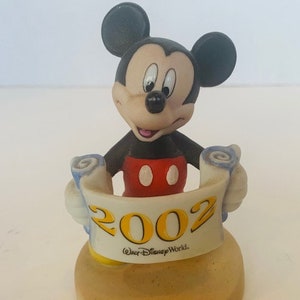 May include: A ceramic figurine of Mickey Mouse holding a scroll with the year 2002 and the text "Walt Disney World." Mickey is wearing his signature red shorts and yellow shoes.