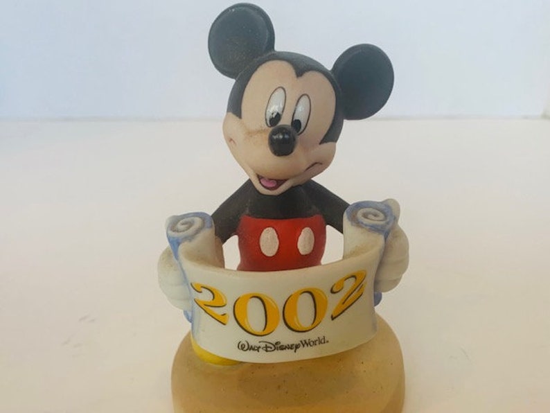 May include: A ceramic figurine of Mickey Mouse holding a scroll with the year 2002 and the text "Walt Disney World." Mickey is wearing his signature red shorts and yellow shoes.