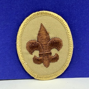 May include: A brown embroidered fleur-de-lis patch on a beige oval background with a yellow border.