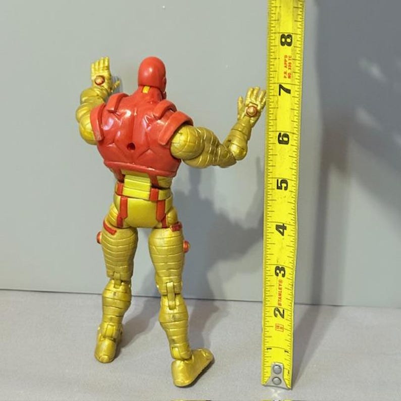 Op de afbeelding: Een gouden en rode action figure van een superheld die op een wit oppervlak staat. De figuur is ongeveer 15 cm hoog.