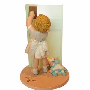 Könnte beinhalten: Eine Porzellanfigur eines jungen Mädchens in einem weißen Kleid mit einer blauen Schleife, das nach einer Tür greift. Eine kleinere Puppe liegt neben ihr auf dem Boden. Der Sockel der Figur ist braun und trägt den Namen der Künstlerin, Bessie Pease Gutmann.