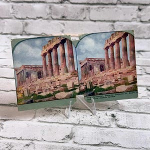 Puede incluir: Una tarjeta estereoscópica vintage que presenta una imagen coloreada del Partenón en Atenas, Grecia. La tarjeta muestra dos fotografías idénticas lado a lado de las antiguas columnas de piedra y ruinas, con el texto The Parthenon, showing Columns on South and Ruins, Athens, Greece impreso en la esquina inferior derecha.