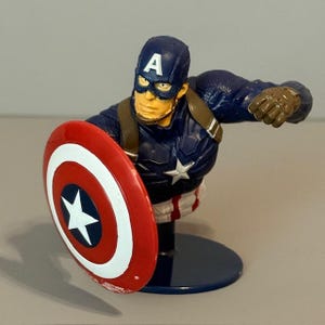 May include: A blue and red Captain America action figure with a shield. The figure is holding the shield in his left hand and has his right arm extended. The figure is standing on a blue base.