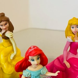 May include: Three Disney princess figurines: Belle in a yellow dress, Ariel in a blue dress, and Aurora in a pink dress.