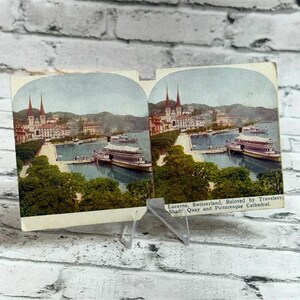Puede incluir: Una tarjeta estereoscópica antigua con dos imágenes coloreadas idénticas de Lucerna, Suiza, que muestran un puerto con un barco de pasajeros, árboles y edificios. El texto dice: Lucerne, Switzerland, Beloved by Travelers, Shady Quay and Picturesque Cathedral.