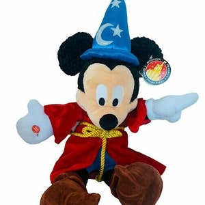 May include: A plush Mickey Mouse wearing a blue wizard hat with a silver crescent moon and star. He is wearing a red robe with gold rope trim and brown shoes. The plush is labeled "Authentic Disney Theme Parks Merchandise."