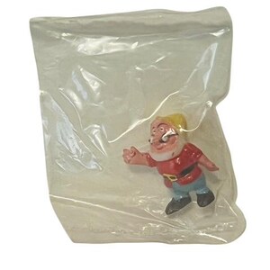May include: A plastic figurine of a dwarf from the Disney film Snow White and the Seven Dwarfs. The dwarf is wearing a red shirt, blue trousers, and a yellow hat. He has a white beard and a moustache.