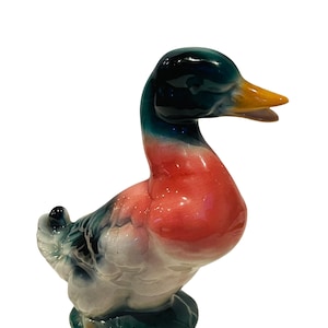 May include: A ceramic duck figurine with a green, pink, and white glaze. The duck has a long neck and a wide beak.