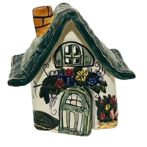 May include: A small ceramic cottage with a green roof, white walls, and a green door. The cottage is decorated with flowers and vines.