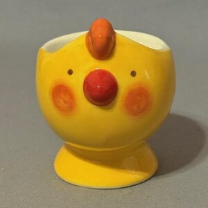May include: A whimsical, yellow ceramic egg cup with a cartoonish face. It features a red nose, orange cheeks, and a small orange crest. The egg cup has a rounded body and a pedestal base.