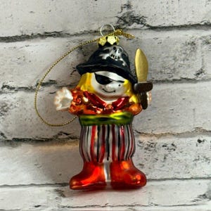 Può includere: Un ornamento pirata in vetro con un cappello nero decorato da un teschio bianco, una benda nera sull'occhio, una sciarpa rossa, una fascia verde in vita e pantaloni a righe rosse e bianche. La figura impugna una spada dorata e ha un gancio dorato.