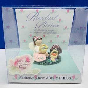 May include: A small, white angel figurine with a pink dress and a basket of flowers. The figurine is sitting on a green base with a pink flower and the words "Rosebud Babies" and "An itty bitty angel to brighten up your day..."  The figurine is in a clear plastic box with a pink and white background. The box also has the words "Love You Eggs-tra Much" and "Exclusively from ABBEY PRESS".